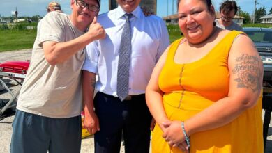 Three people posing outdoors on a sunny day: a man in a white shirt and tie in the center, a man in a beige shirt giving a thumbs up on the left, and a woman in a bright yellow dress on the right.