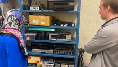 Two people stand at a blue metal shelving unit filled with electronics, examining vintage audio gear and printers on multiple shelves.