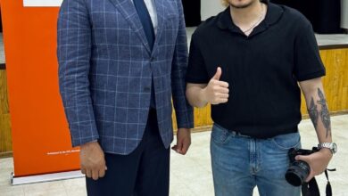Two men pose for a photo indoors: a man in a blue checkered suit beside a smiling photographer holding a camera, with an orange Manitoba NDP banner in the background.