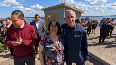 Group of people posing for a photo outdoors on a sunny day near a small wooden shed by a body of water; smiles and casual attire.