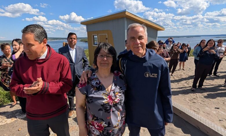 Group of people posing for a photo outdoors on a sunny day near a small wooden shed by a body of water; smiles and casual attire.