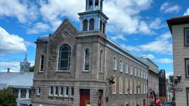Brick building with a tall clock tower and arched windows along a city street; pedestrians nearby under a bright blue sky with clouds.