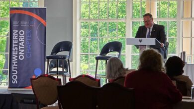 Male speaker in a suit at a lectern delivering a talk in a bright, window-filled conference room with a blue banner to the left.