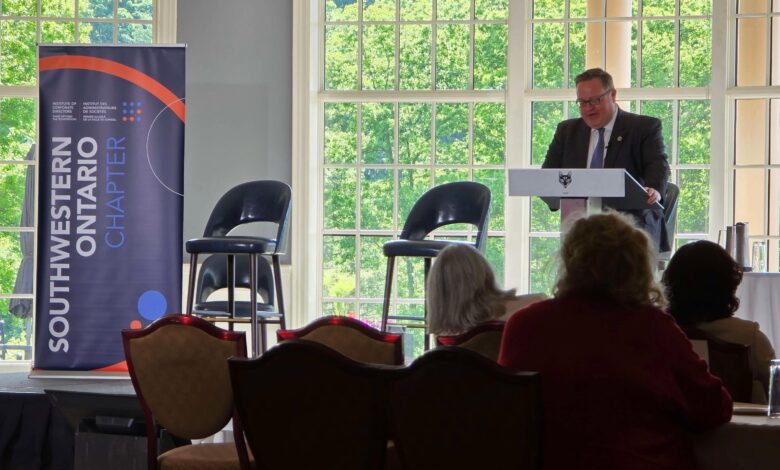 Male speaker in a suit at a lectern delivering a talk in a bright, window-filled conference room with a blue banner to the left.