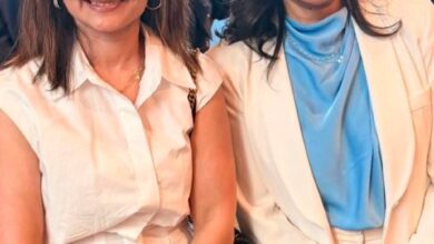 Two women posing at a formal event; left in a white blouse and black skirt, right in a white suit with a light blue top.