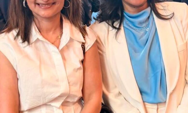 Two women posing at a formal event; left in a white blouse and black skirt, right in a white suit with a light blue top.