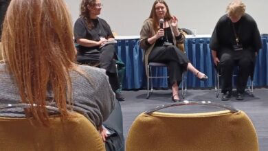 Panel discussion with four women seated on a stage; the center speaker holds a microphone and talks as the others listen.