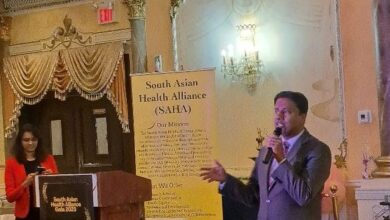 Speaker in a gray suit with a microphone at a podium during a SAHA event; a yellow banner behind him reads 'South Asian Health Alliance (SAHA)'.