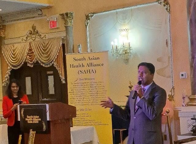 Speaker in a gray suit with a microphone at a podium during a SAHA event; a yellow banner behind him reads 'South Asian Health Alliance (SAHA)'.