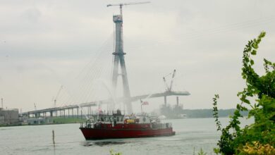 Red tugboat on a calm river with a towering crane and a partially built bridge in a hazy sky.