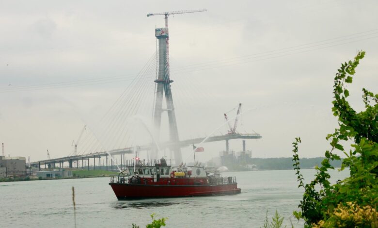 Red tugboat on a calm river with a towering crane and a partially built bridge in a hazy sky.