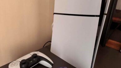 PlayStation 5 console standing upright on a table with two controllers—white and purple DualSense—in front of it.