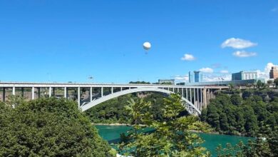White hot air balloon drifting over a turquoise river with an arched bridge and city skyline in the distance under a clear blue sky.