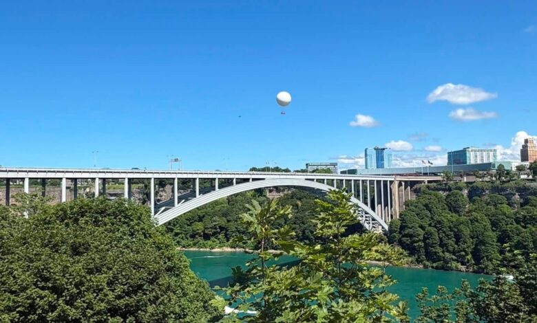 White hot air balloon drifting over a turquoise river with an arched bridge and city skyline in the distance under a clear blue sky.