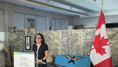 Woman in a black dress speaks at a podium during a family-violence prevention event, with a Canadian flag to her right and a banner on the lectern reading 'Preventing family violence' (English and French).