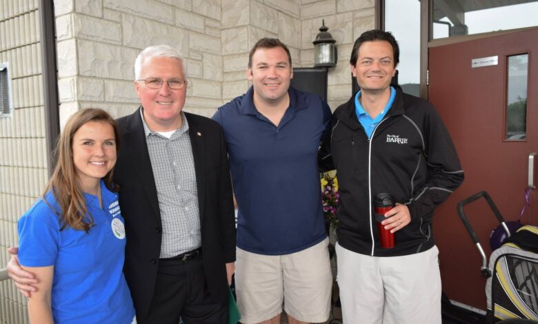 Four adults posing outside a stone building, smiling at the camera; a woman in a blue polo on the left, and three men in smart casual attire.