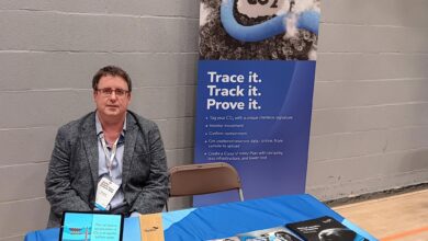 Man in a gray blazer seated at a booth with CO2-themed banners and brochures on a blue tablecover, at an indoor expo setting.