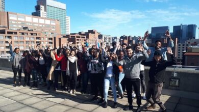 Diverse group on a rooftop, raising hands in the air, with a city skyline and blue sky behind them.