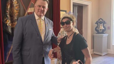 Man in a blue suit with a yellow striped tie stands beside a woman wearing a tiara and large sunglasses at an event location with red curtains in the background.
