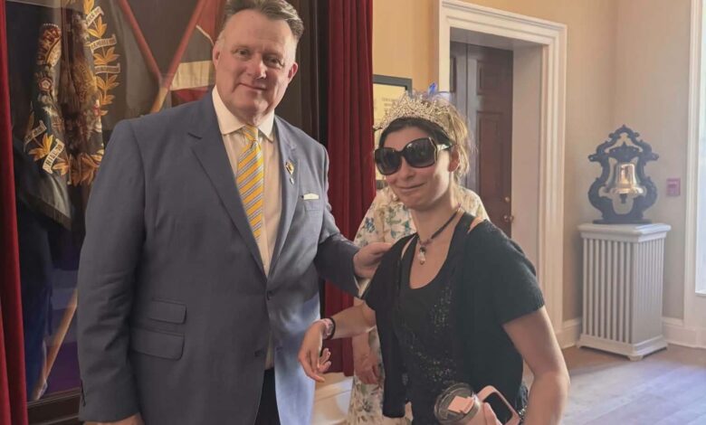 Man in a blue suit with a yellow striped tie stands beside a woman wearing a tiara and large sunglasses at an event location with red curtains in the background.