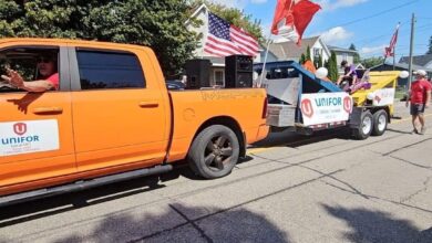 Orange pickup truck towing a parade float with UNIFOR banners and a Canadian flag, along a sunny residential street with spectators and other floats in the background.