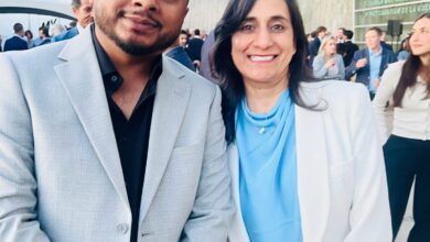 Two adults posing at an outdoor event; man in a light gray suit and black shirt, woman in a white suit with a blue blouse, with a museum exterior in the background.