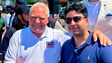 Two men shake hands outdoors, one in a white Ford Nation polo and the other in a blue polo, with storefronts behind him.